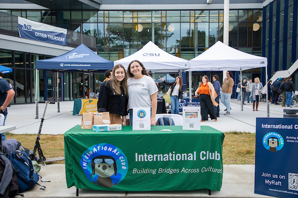 two students posing and smiling in front of a table for the International Otters Club
