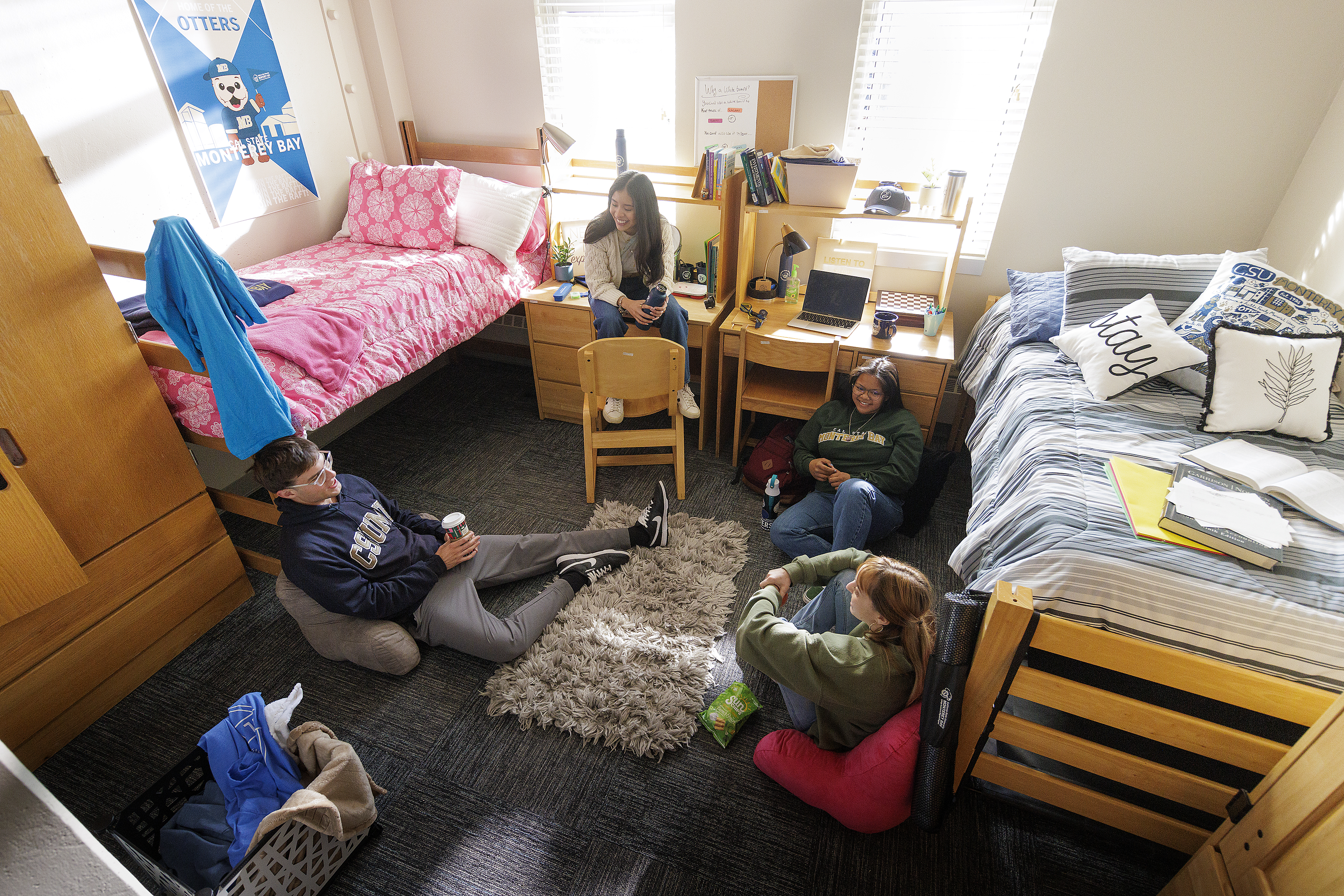 4 students in circle in dorm room talking, two students on the ground, one student on the bed, and one student sitting on a chair