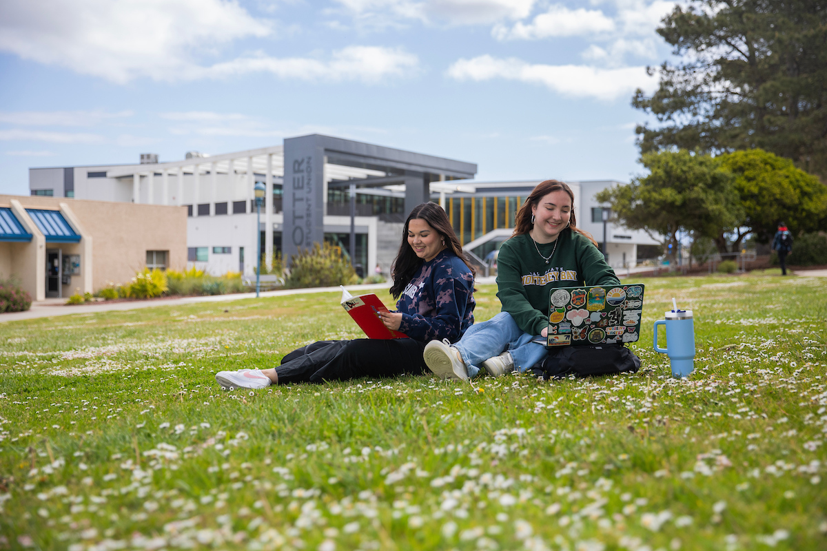 Two CSUMB students sitting on the grass in Main Quad with the Otter Student Union (OSU) in the background